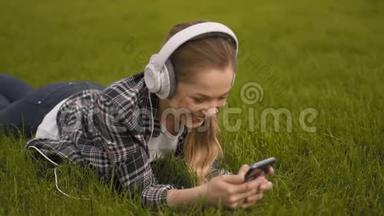 一个年轻的女人戴着大耳机躺在草地上，在电话上看视频。 搜索、网络搜索4Kk